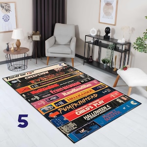 May include: Rectangular area rug featuring a design of stacked VHS tapes. The rug displays titles like "Friday the 13th," "Child's Play," and "Halloween 4." The rug is primarily black with colorful text and imagery.