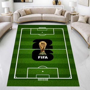 May include: Green rectangular area rug with a soccer field design. The rug features white lines marking the field, a large black number 26, and a gold trophy with the word "FIFA" at the bottom. The rug is placed in a living room with beige sofas and white walls.