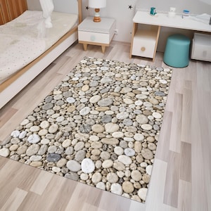 Puede incluir: Una alfombra rectangular con un estampado fotográfico de alta resolución de piedras de río lisas y redondeadas en varios tonos de gris, beige, blanco y bronceado. El patrón crea una textura realista de una superficie cubierta de guijarros.