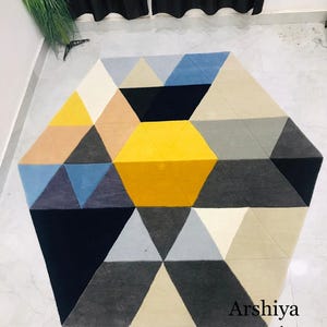 May include: Geometric area rug with a hexagonal shape, featuring a pattern of triangles in various colors including yellow, blue, black, gray, and beige. The rug has the text "Arshiya carpets" in the bottom right corner.