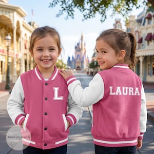 May include: A pink and white varsity jacket featuring a white letter L on the front left chest and the name LAURA in white block letters on the back. The jacket has a snap-button closure, white sleeves, and striped ribbed trim on the collar and cuffs.