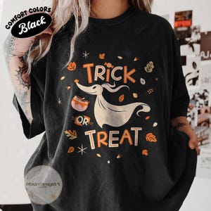 May include: A black short-sleeve t-shirt featuring a graphic of a ghost dog and the text "TRICK OR TREAT" in orange and cream colors. The design includes scattered autumn leaves, small stars, and a candy bowl, set against a dark fabric background.
