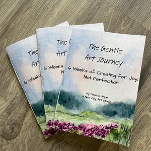 May include: Three softcover books titled "The Gentle Art Journey" are displayed. Each book features a watercolor design with a blue sky, green hills, and purple flowers. The text includes "6 Weeks of Creating for Joy, Not Perfection."