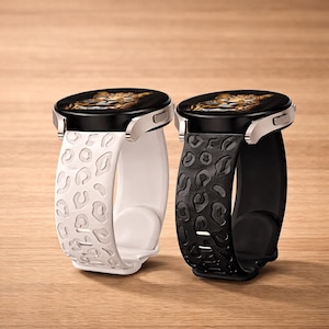 Puede incluir: Dos smartwatches con pantallas negras y biseles plateados. Uno tiene una correa blanca con un diseño de estampado de leopardo, y el otro tiene una correa negra con un patrón similar. Los relojes se muestran sobre una superficie de madera.