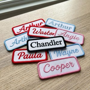May include: A collection of rectangular embroidered name patches with white fabric centers and colored borders. The patches feature names including Arthur, Wastor, Angie, Chandler, Paula, Wayne, and Cooper stitched in various fonts and thread colors.