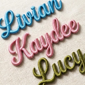May include: Three embroidered name patches in cursive script. The top patch is light blue and reads Livian. The middle patch is light pink and reads Kaydee. The bottom patch is olive green and reads Lucy. Each patch features a raised, satin-stitched texture.