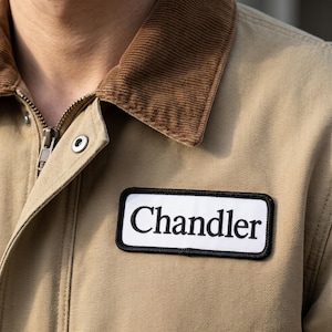 May include: A tan canvas jacket featuring a brown corduroy collar, a metal zipper, and a rectangular white patch on the chest with a black border and the word Chandler embroidered in black serif lettering across the center of the patch.