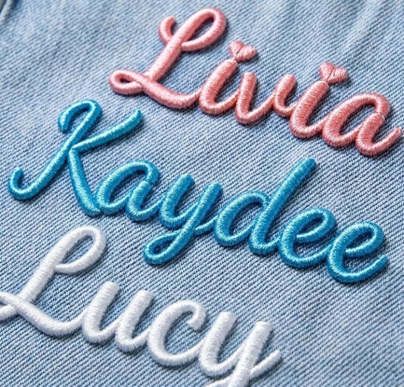 May include: Light blue denim fabric featuring three rows of embroidered names in cursive script. The top name Livia is stitched in pink thread, the middle name Kaydee is in bright blue thread, and the bottom name Lucy is in white thread.