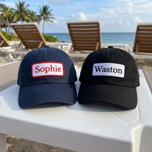 Puede incluir: Una gorra de béisbol azul marino con un parche rectangular que muestra el texto rojo Sophie y una gorra de béisbol negra con un parche rectangular que muestra el texto negro Waston. Ambas gorras tienen una visera curva.