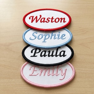 May include: Four oval-shaped embroidered patches with white fabric centers. Each patch features a name stitched in a different color: Waston in red, Sophie in light blue, Paula in black, and Emily in pink. Each patch has a matching colored border.
