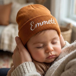 Puede incluir: Un gorro de punto acanalado de color óxido con el nombre Emmett bordado en letras cursivas blancas en el dobladillo frontal. El gorro presenta un borde doblado y un patrón de punto texturizado, ofreciendo una prenda sencilla y funcional.