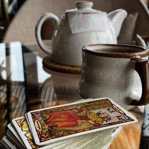 Può includere: Un mazzo di carte dei tarocchi con un'illustrazione colorata di una regina in abito rosso. Una teiera in ceramica bianca e una tazza con manico e bordo marrone sullo sfondo. Gli oggetti sono su un tavolo di vetro.