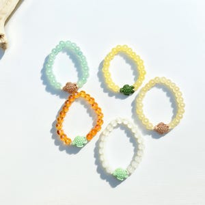 May include: Five beaded bracelets in shades of green, yellow, orange, and white, each featuring a turtle charm. The bracelets are arranged on a white surface, with a piece of driftwood in the upper left corner. The beads appear to be approximately 0.25 inches in diameter.