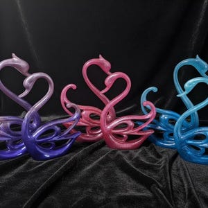 May include: A collection of decorative swan sculptures in shades of purple, pink, and blue. The swans are stylized with intertwined necks forming a heart shape. The sculptures have a glossy finish and are arranged on a dark, textured surface.