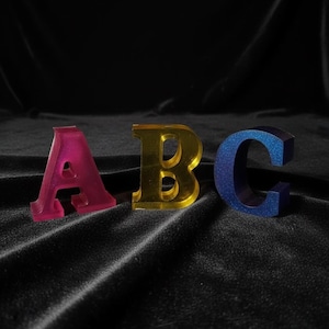 May include: Three-dimensional letters A, B, and C in a row. The letter A is a translucent pink, B is a shiny gold, and C is a glittery blue. The letters are displayed on a black, textured surface.