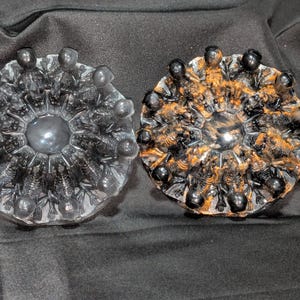 May include: Two circular resin coasters featuring a radial arrangement of small, molded skeleton figures. The left coaster is translucent with black skeletons, while the right coaster features black skeletons with orange highlights on a dark background.