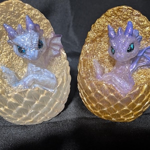 May include: Two small dragon figurines emerging from textured, egg-shaped shells. The left dragon is iridescent white with blue eyes, while the right dragon is translucent purple with dark eyes. Both eggs feature a gold-toned, scaly exterior finish.