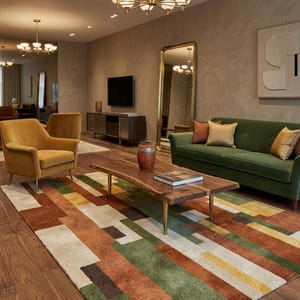 May include: A modern living room with a green sofa, mustard-colored armchair, and a wooden coffee table. A colorful geometric rug covers the hardwood floor. The room is illuminated by a brass chandelier and recessed lighting.