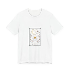 May include: White t-shirt featuring a tarot card design. The design includes a crescent moon, sun, hands, and floral elements. The text "THE INTUITIVE" is at the bottom of the card.