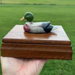 May include: A rectangular wooden box with a smooth, polished finish, featuring a small, hand-painted ceramic mallard duck figurine attached to the center of the lid. The duck has a green head, a yellow beak, a reddish-brown chest, and grey wings.
