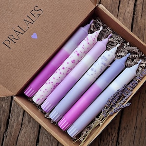 Puede incluir: Una caja con seis velas cónicas moradas con diseños variados, incluyendo patrones moteados y a rayas. La caja está etiquetada como "PRÁLAIES" y contiene ramitas de lavanda seca. Las velas están dispuestas en una caja de cartón marrón.