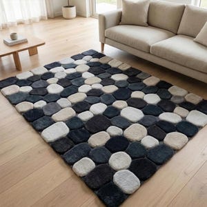 May include: A square area rug with a pebble design in shades of gray, black, and beige. The rug is made of a soft, textured material and is placed on a light-colored wooden floor. The rug is in a living room setting.