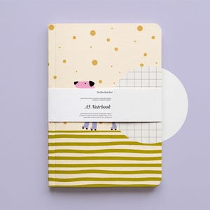 Op de afbeelding: A5-notitieboek met een crèmekleurige kaft met gouden stippen en een cartoonhondenillustratie. De onderste helft heeft olijfgroene horizontale strepen. Een witte band wikkelt zich om het notitieboek met de tekst "The New First Book" en "A5 Notebook".