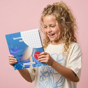 May include: A young girl smiles while holding a children's storybook titled "Secrets the". The book cover features a blue underwater scene with illustrations of fish. The girl wears a cream-colored t-shirt with a blue floral design.