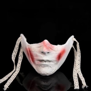 May include: A white fabric mask with red paint around the nose and mouth, and cream lace ties. The mask has a distressed look, suggesting a horror or Halloween theme. The background is black.