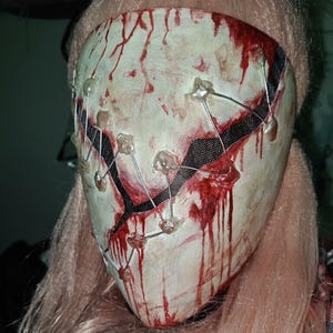 May include: A pale mask with red streaks and drips, resembling blood. The mask has a stitched appearance with wire and clear, rounded fasteners. A black mesh section is visible. The mask is set against a background of pink hair.