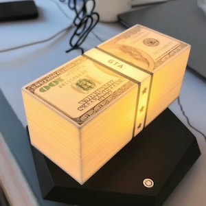 以下が含まれることがあります： 100ドル札の束を模した長方形のランプで、バンドには「GTA」の文字が入っています。 ランプは暖かく魅力的な光を放ち、電源ボタンが付いた黒いベースの上に置かれています。