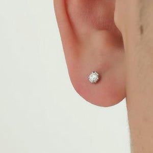 May include: A close-up of a person's ear with a small, round, diamond stud earring. The earring is set in a silver or white metal and sparkles. The skin around the ear is light-toned, and the background is a soft, neutral color.
