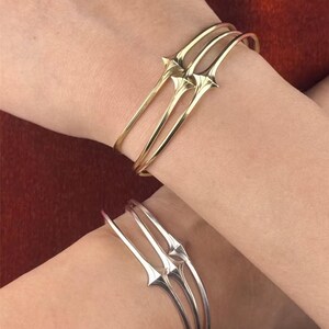 Può includere: Due braccialetti color oro e argento. Ogni braccialetto presenta tre sottili fasce con un design stilizzato e appuntito al centro. I braccialetti sono indossati sui polsi, mostrando il loro design minimalista ed elegante.