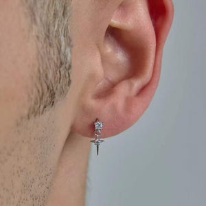 May include: A silver-toned stud earring featuring a small round clear gemstone at the top, connected to a dangling four-pointed star charm with a tiny clear gemstone at its centre. The earring is secured through the earlobe with a metal post.