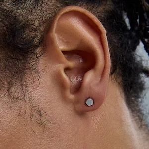 Peut inclure: Gros plan sur une oreille avec une petite boucle d'oreille hexagonale argentée. La boucle d'oreille est dans le lobe de l'oreille. La peau est de couleur moyenne et les cheveux sont foncés et bouclés. La boucle mesure environ 0,5 cm.