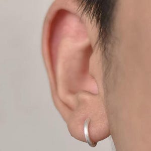 以下が含まれることがあります： 耳たぶに装着された、マットでブラッシュ仕上げの小さな円形のシルバーフープピアス。ピアスはシンプルな連続したバンドデザインで、控えめな外観を作り出しています。金属は柔らかく、光沢のない質感をしています。