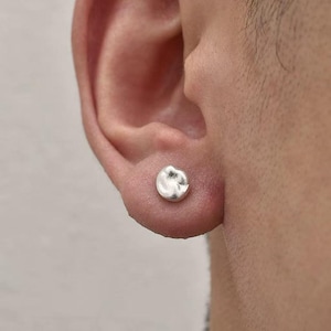 May include: A small, silver stud earring with a textured, organic design. The earring is round and sits on the earlobe. The background is a blurred view of a person's ear and face.