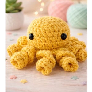 May include: A handmade, yellow crochet octopus with black button eyes. The octopus has eight curled tentacles and is sitting on a light-colored wooden surface. Decorative elements include a small succulent plant and colorful yarn balls.