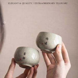 Może przedstawiać: Dwie jasnozielone filiżanki do herbaty z krakelą na glazurze trzymane są w dłoniach. Filiżanki mają zaokrąglone kształty i chińskie znaki. U góry znajduje się napis "GE KILN MASTER TEA CUP" i "ELEGANT & QUALITY / EXTRAORDINARY TEAWARE".