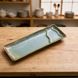 Może przedstawiać: Prostokątny ceramiczny talerz do serwowania z jasnoniebieskim i zielonym szkliwem. Talerz ma teksturowaną krawędź i falisty separator. Ręcznie robione naczynie stoi na drewnianej powierzchni.