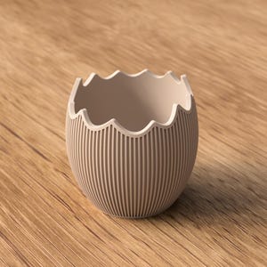 Op de afbeelding: Een beige eivormige decoratieve container met een geschulpte rand en verticaal gestreept patroon. De binnenkant is lichtbeige. De container staat op een houten oppervlak.