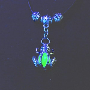 Peut inclure: Un collier en argent avec un pendentif grenouille vert qui brille dans le noir. La grenouille est détaillée avec un contour argenté et a un corps rond et vert.