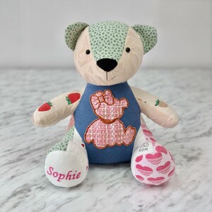 Pode incluir: Um urso de pelúcia feito à mão com um design de patchwork. O urso apresenta um colete jeans azul com uma aplicação de urso rosa, braços e orelhas estampados com morangos e flores, e uma perna com corações rosa. O nome "Sophie" é bordado em um pé.