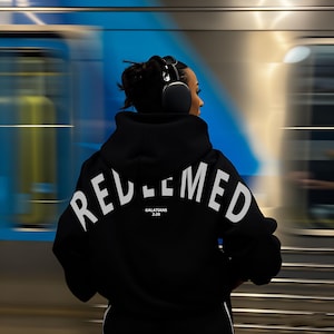 May include: A black hoodie with the word "REDEEMED" in white arched across the back. Below the word is the text "GALATIANS 2:20". The person in the image is wearing black headphones.