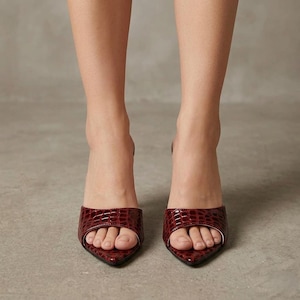 May include: A pair of burgundy-colored, open-toe mules with a pointed toe and a textured, crocodile-like pattern. The shoes have a small heel and are shown from a front-facing perspective.