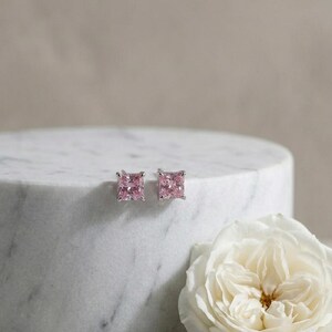 May include: A pair of square-cut pink gemstone stud earrings. The earrings are set in a silver metal and are displayed on a white marble surface, next to a white rose. The earrings are a classic jewellery item.