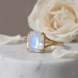 May include: A gold ring featuring a large, cushion-cut moonstone with a translucent, iridescent blue glow. The gemstone is held by four prongs on a polished gold band that splits into a dual-shank design as it reaches the setting.