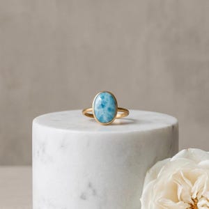 Könnte beinhalten: Ein goldener Ring mit einem großen, oval geformten, hellblauen Edelstein. Der Edelstein hat ein marmoriertes Aussehen mit weißen Einschlüssen. Der Ring liegt auf einer weißen Marmoroberfläche, mit einer weißen Rose im Hintergrund.