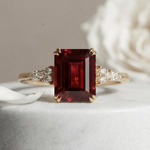 May include: A gold ring featuring a large, rectangular, emerald-cut deep red gemstone at the centre. The band is accented on both sides by a cluster of three small, round, clear white diamonds set in a triangular arrangement on the gold shank.