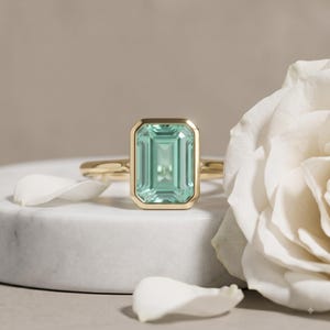 Puede incluir: Un anillo de oro con una gran piedra preciosa de color verde menta, de corte esmeralda rectangular y engastada en un bisel liso. El anillo está colocado en el centro, mostrando la piedra facetada clara y la banda de oro amarillo pulido.